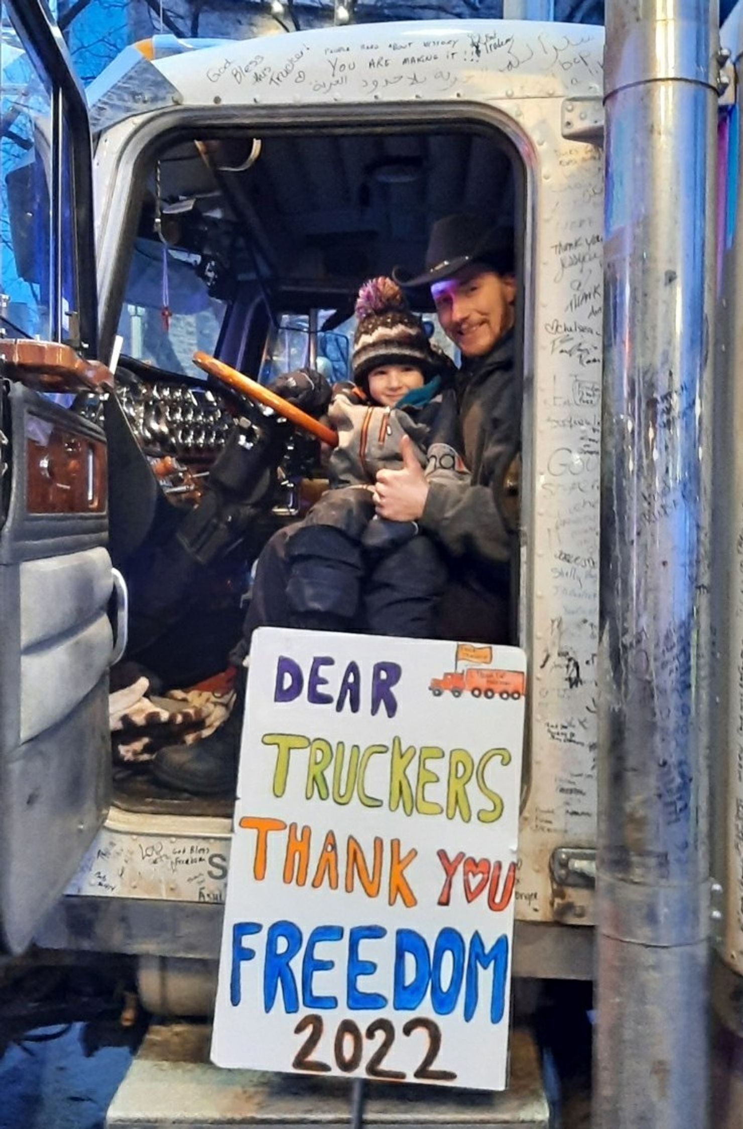 Image of Spencer Pretty Boy Bautz sitting in the drivers seat of a semi truck with a child sitting on his lap